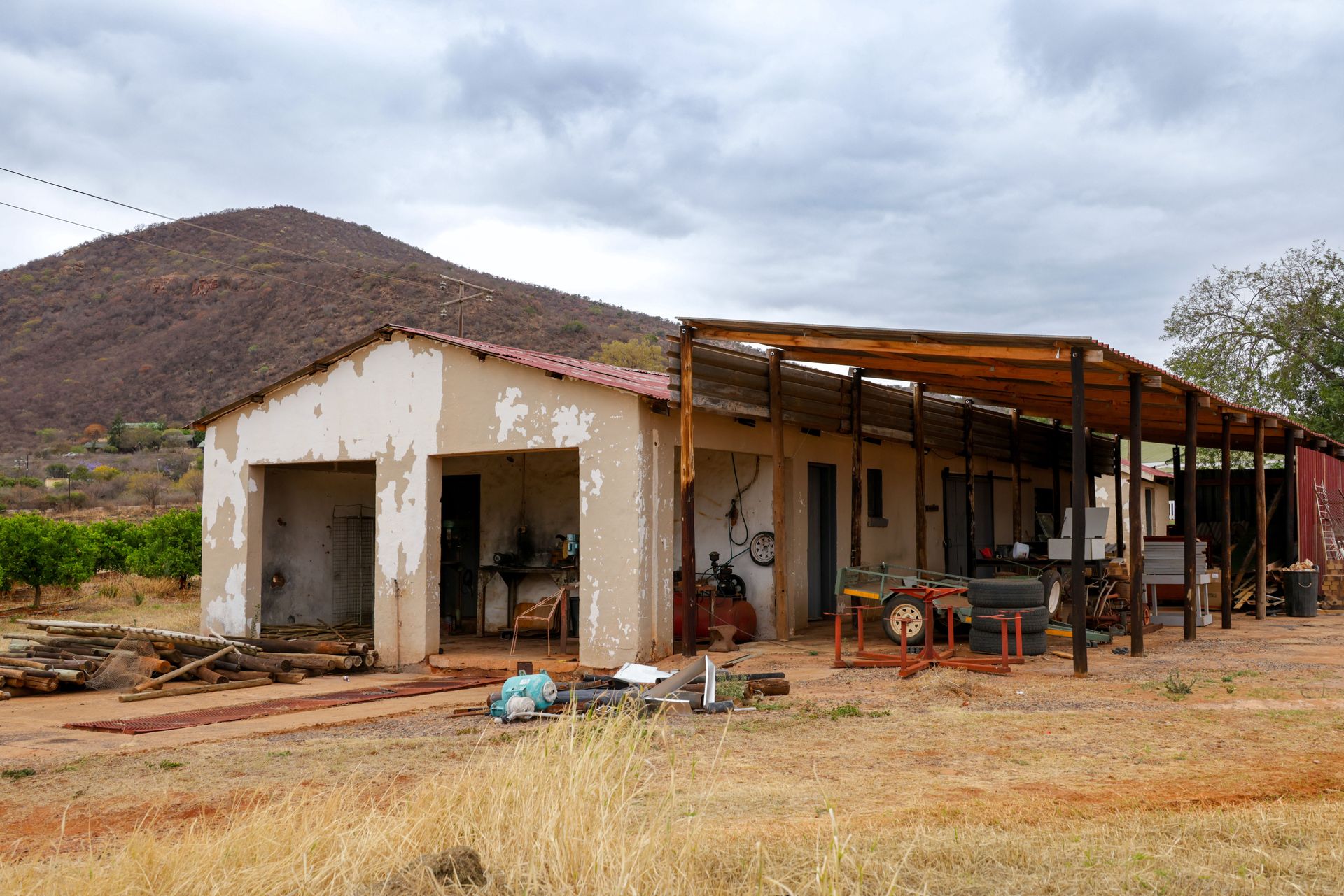 Rustic farmyard with a workshop and mountains in the background under cloudy skies.